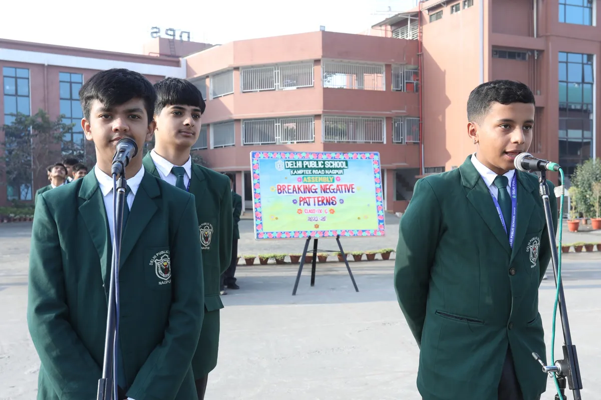 DPS (Kamptee Road) students in green blazers speaking into microphones at an outdoor school assembly, with a decorated assembly board and school building behind them.