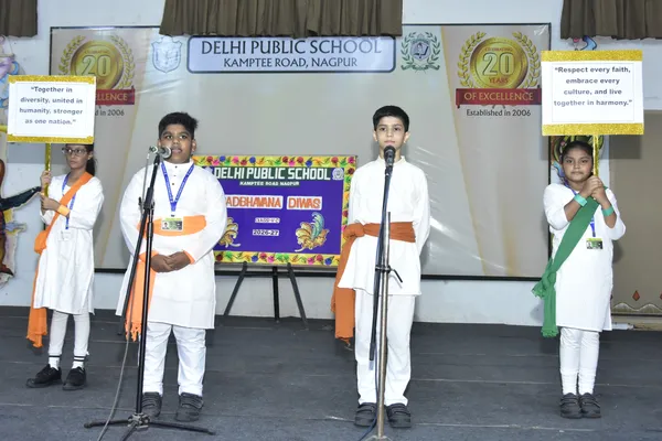 Four students in white and saffron outfits stand on a stage at DPS (Kamptee Road), two at microphones and two holding placards, in front of a school banner and backdrop.