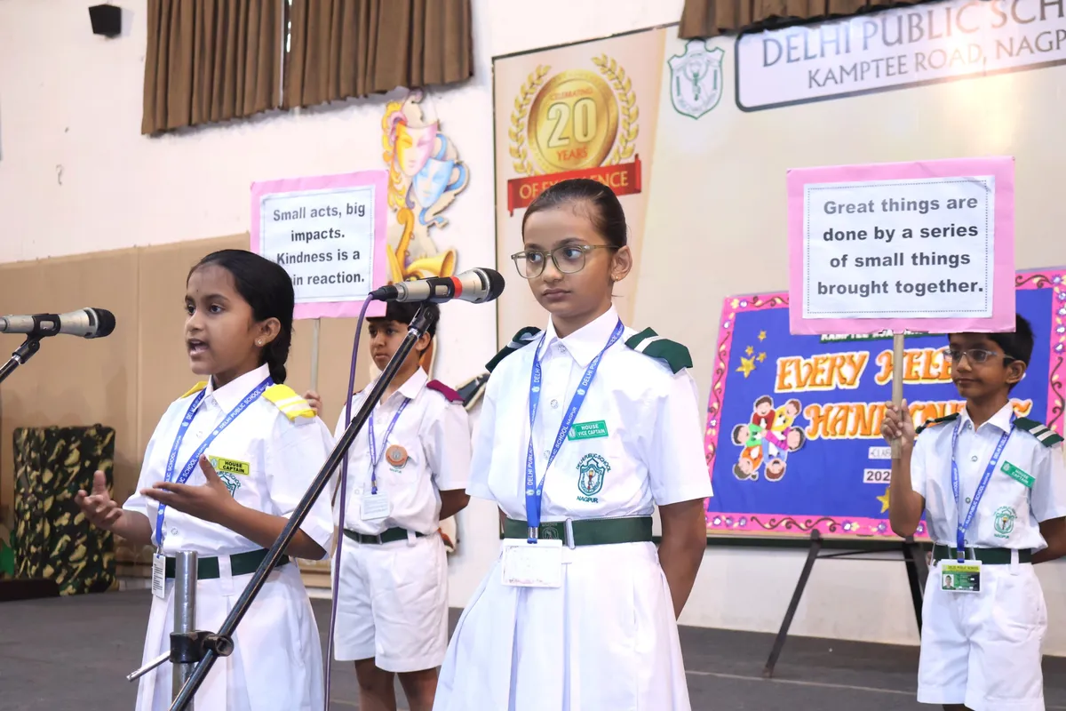 DPS (Kamptee Road) students in school uniforms speaking at microphones on a stage, holding small placards with messages, with decorated boards behind them.