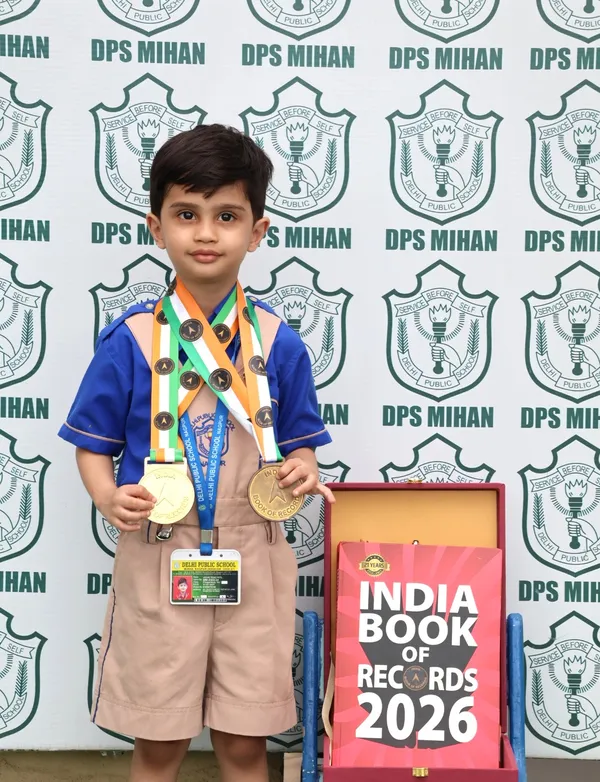 A young boy in school uniform wears several medals and stands beside a boxed record book in front of a DPS (Mihan) branded backdrop.