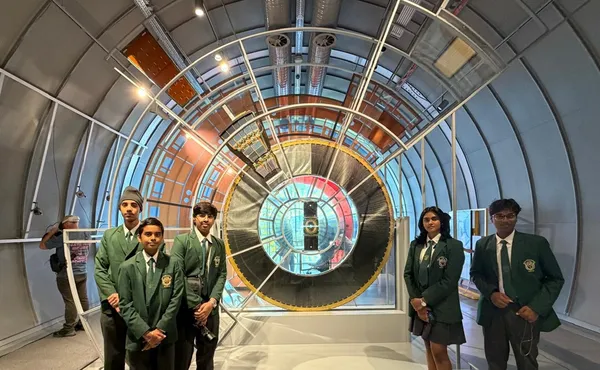Five students in green blazers posing in front of a large circular scientific exhibit inside a curved tunnel-like hall