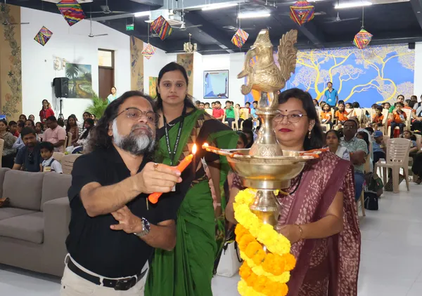 A man and two women light a brass lamp decorated with marigold flowers while an audience watches in a hall hung with paper kites at DPS (Mihan).