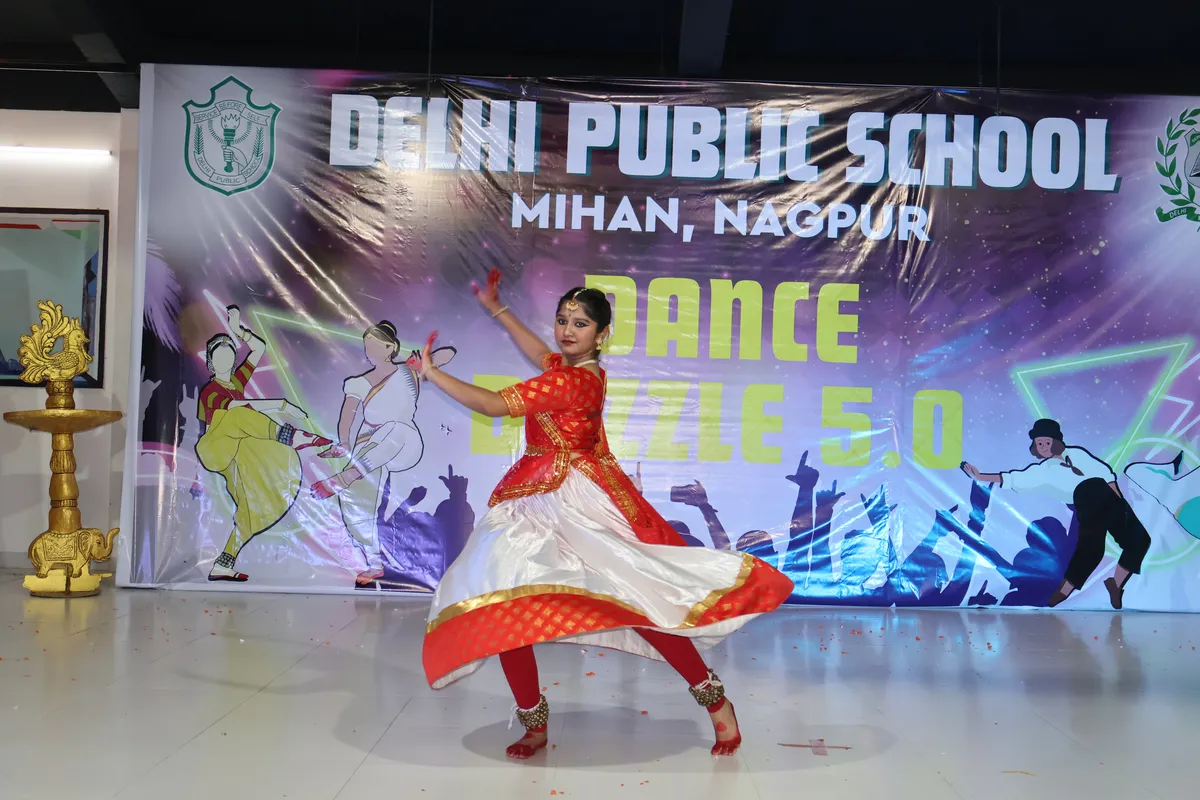 A girl in a red and white classical dance costume performs on a stage in front of a DPS (Mihan) dance event banner.