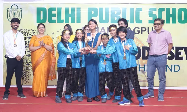 Group of students in blue sparkly outfits holding a trophy and medals on stage with adults standing beside them in front of a school banner. - DPS (Mihan)