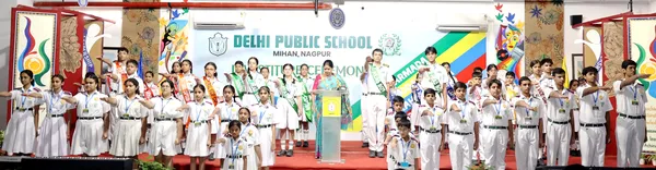 Students in uniform and sashes standing in rows on stage at DPS (Mihan) Investiture Ceremony with woman at podium