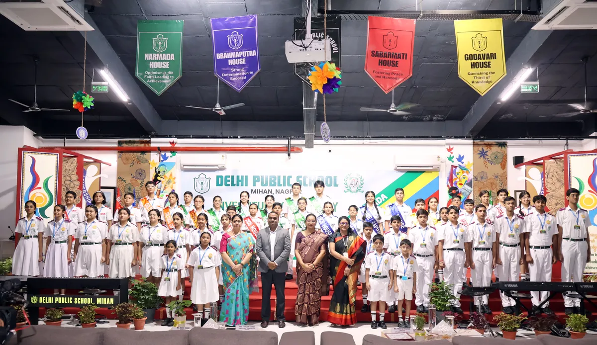 Group photo of students in school uniforms and sashes with adults standing in front of a stage banner at DPS (Mihan) Investiture Ceremony