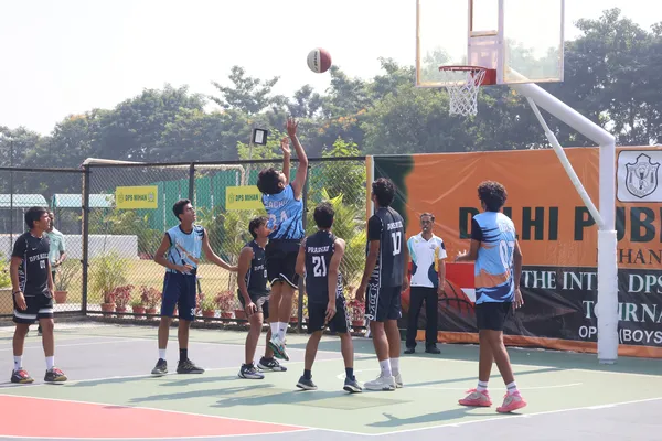 Boys playing basketball on an outdoor court, one jumping to shoot near the hoop while others and a referee watch. - DPS (Mihan)