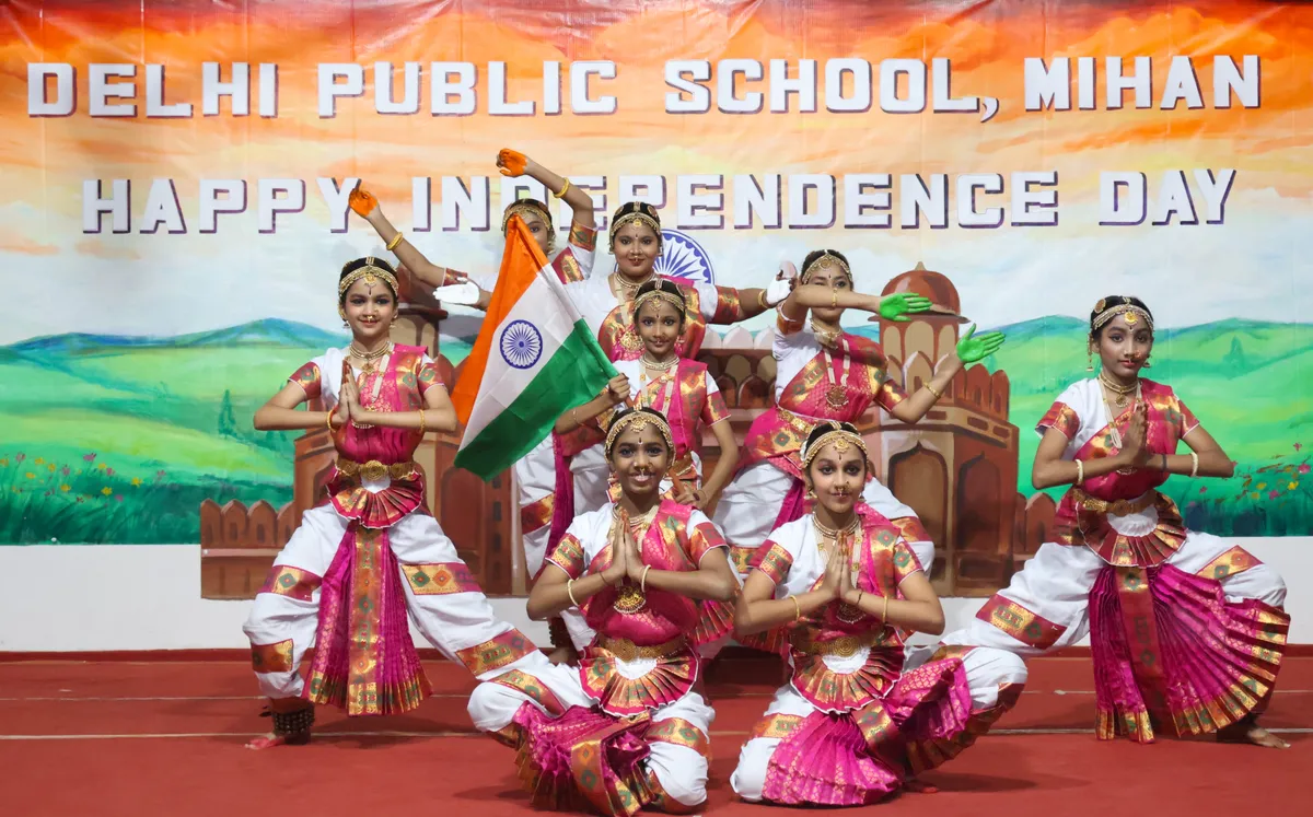 Group of girls in classical dance costumes posing on a stage in front of an Independence Day backdrop with an Indian flag. - DPS (Mihan)