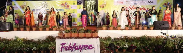 Children in colourful costumes standing on a stage during a fashion show at DPS (Mihan), with banners and potted plants