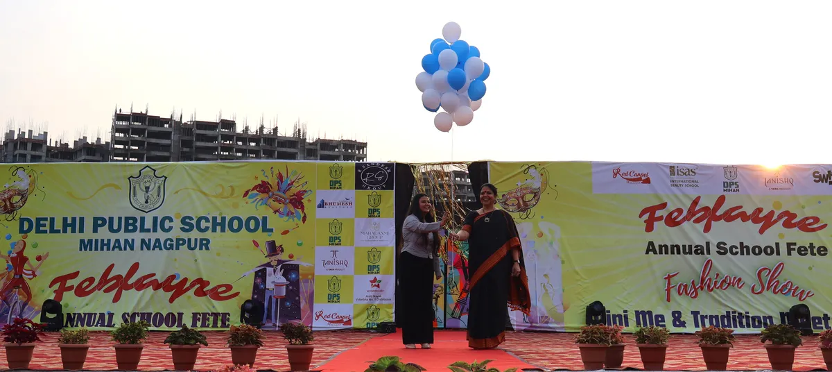 Two women greeting each other on a stage beneath blue and white balloons in front of a DPS (Mihan) school fete banner