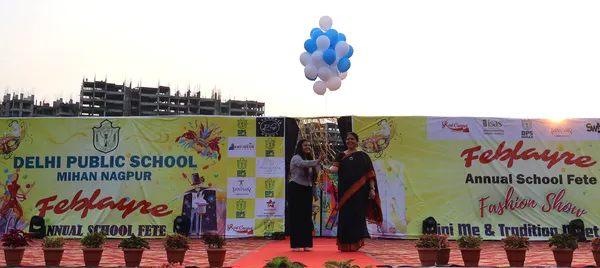 Two women greeting each other on a stage beneath blue and white balloons in front of a DPS (Mihan) school fete banner