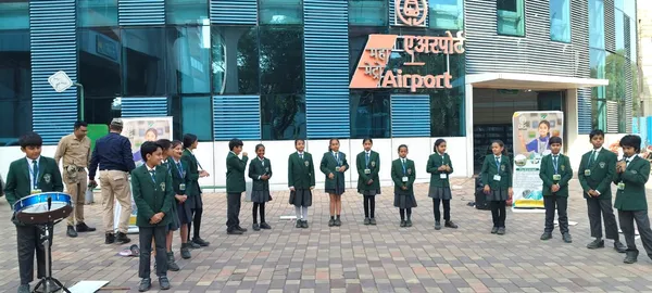 Students in green uniforms stand in a line outdoors near a metro airport station with a drum on the left and two men standing nearby.