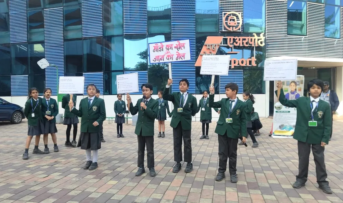Students in green uniforms hold placards and a microphone outdoors in front of a metro airport station building.