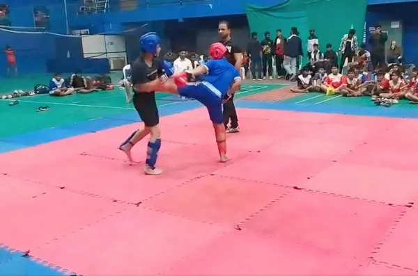 Two DPS (Mihan) athletes in protective gear sparring on a mat, one delivering a high kick, with a referee and seated spectators behind