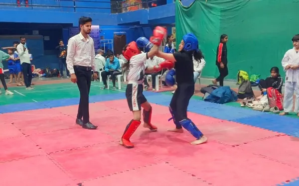 Two DPS (Mihan) students in headgear and gloves sparring on a red mat while a referee in a white shirt watches and others sit nearby