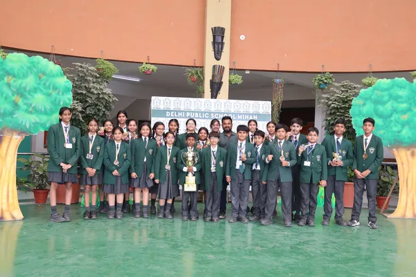 Group photo of about 20 DPS (Mihan) students in green blazers, some wearing medals and one holding a trophy, posing indoors in front of a banner