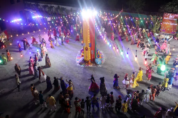 Aerial night view of people in colorful traditional clothes dancing around a lit, decorated central pillar with string lights. - DPS (Mihan)