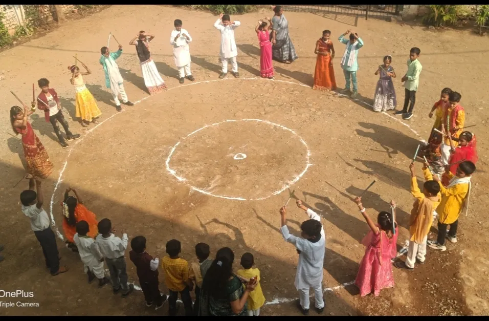 Aerial view of children in colorful traditional clothes holding sticks, standing in a large circle drawn on dirt ground, with a few adults. - Ekatmata Upper Primary School