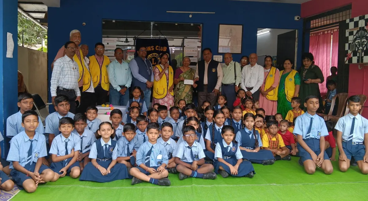 Group photo of many school children in uniforms seated on a green mat with a row of adults, some in yellow vests, standing behind them in a hall. - H.S. Mahatme Sanskar Vihar School