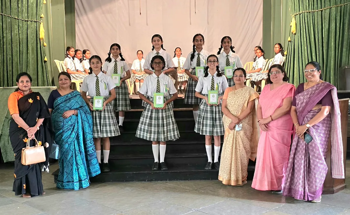 Group photo of schoolgirls in uniform holding green booklets on stage steps, with five women in sarees standing alongside them at J. N Tata Parsi.