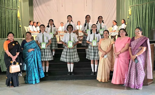Group photo of schoolgirls in uniform holding green booklets on stage steps, with five women in sarees standing alongside them at J. N Tata Parsi.