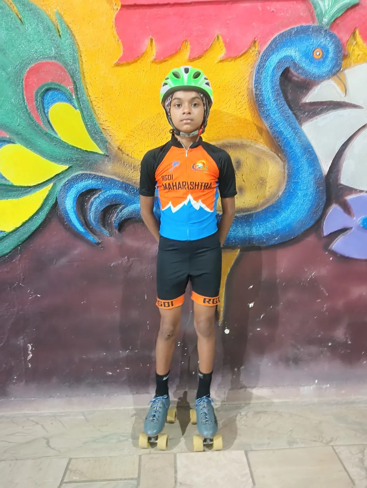 A child in a green helmet, orange and blue jersey and black shorts stands on roller skates in front of a colorful peacock mural. - J. N Tata Parsi