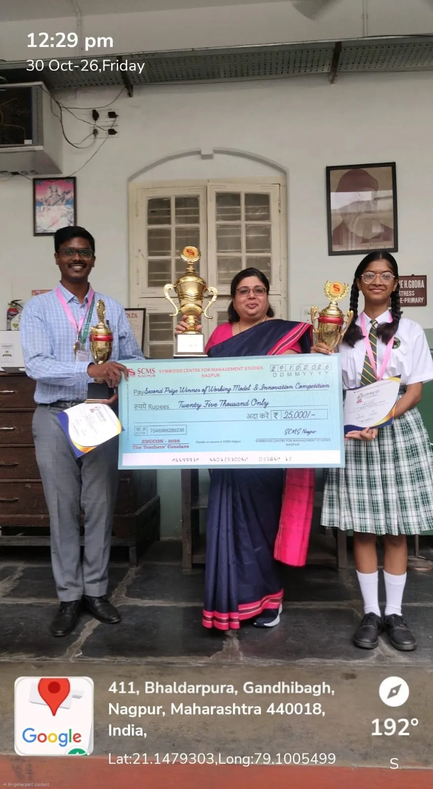 Three people, including a student in school uniform, stand holding trophies and a large prize cheque indoors, with a GPS map overlay at the bottom. - J. N Tata Parsi