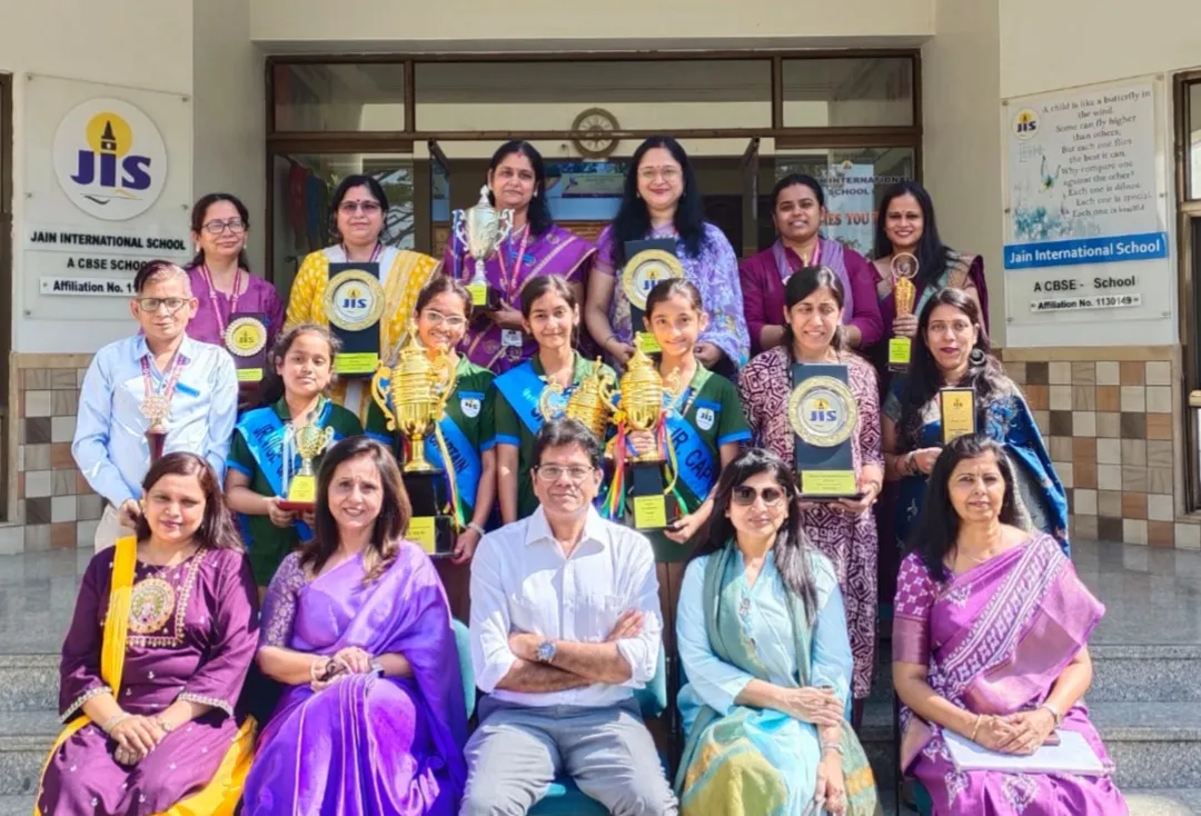 Group photo of teachers and students on school entrance steps holding trophies and plaques, with Jain International school name boards visible behind them.
