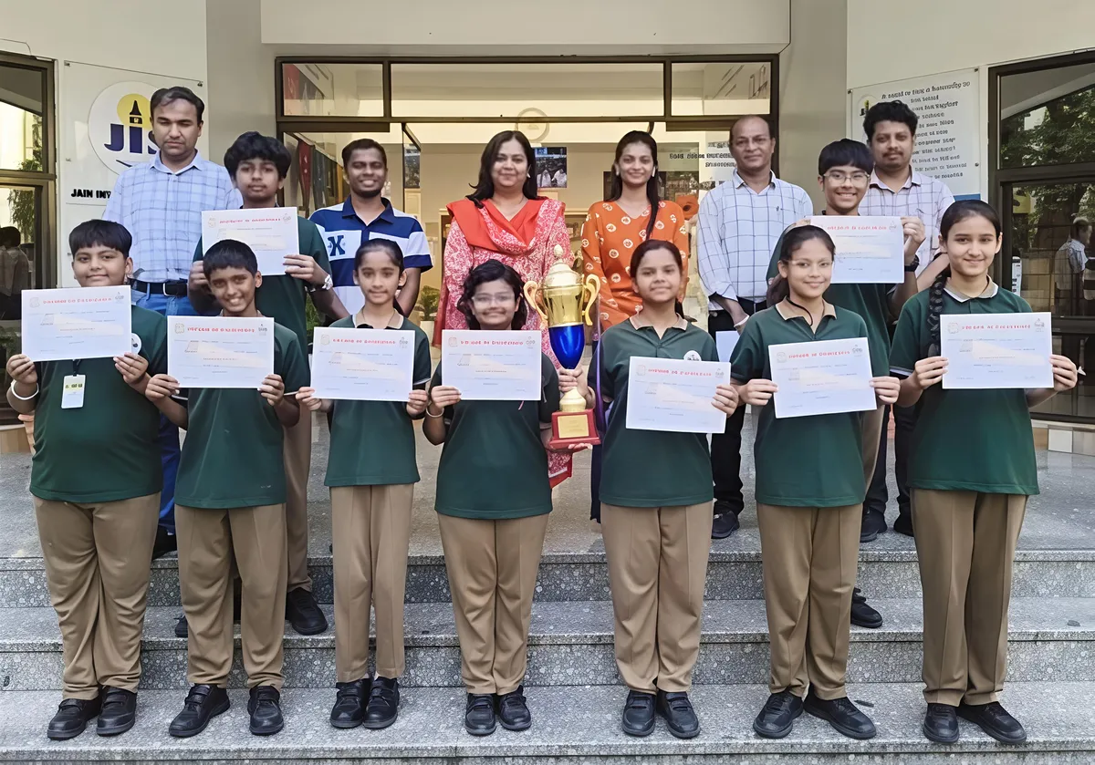 Group photo of students in green uniforms holding certificates and a trophy, standing on steps with teachers behind them outside a school entrance. - Jain International