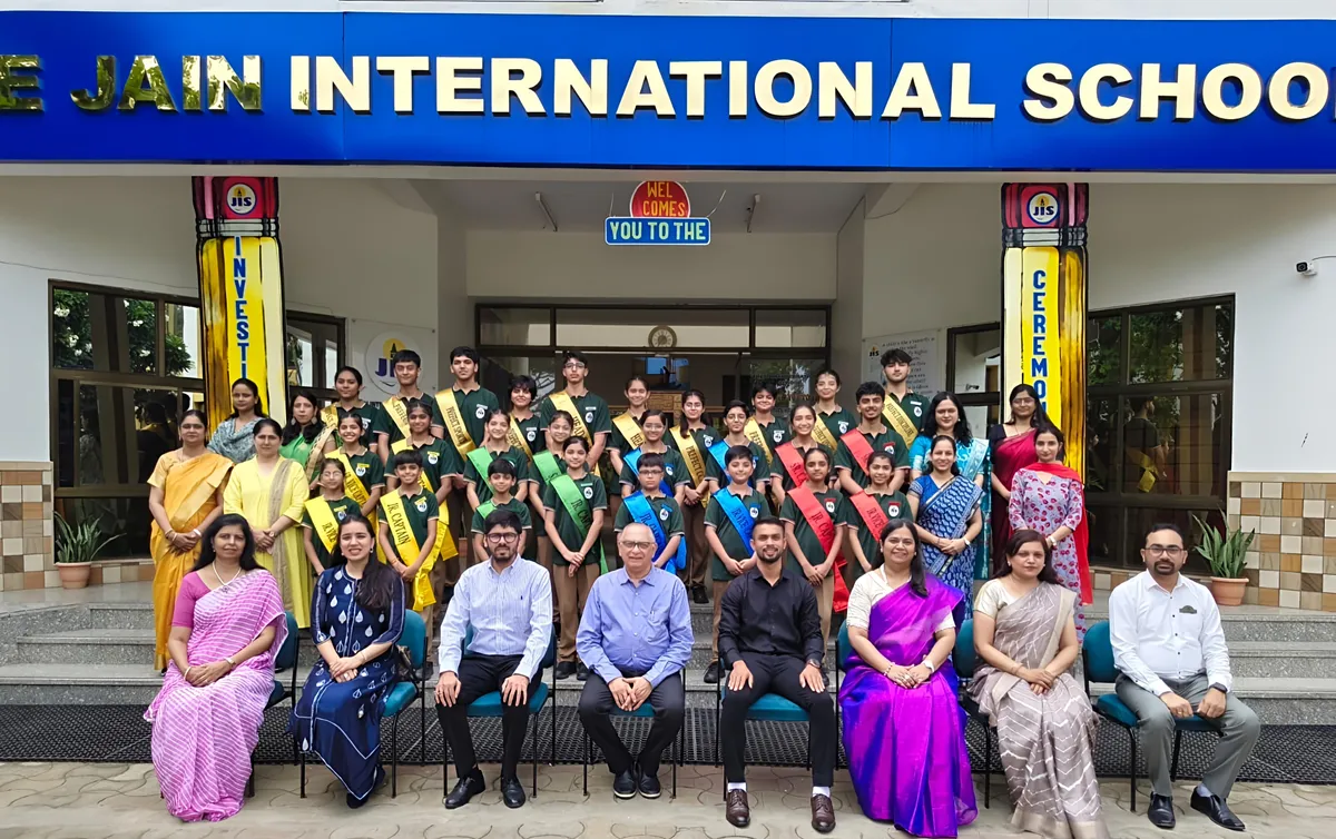 Group photo of students wearing coloured sashes standing with seated teachers and guests in front of Jain International school entrance.