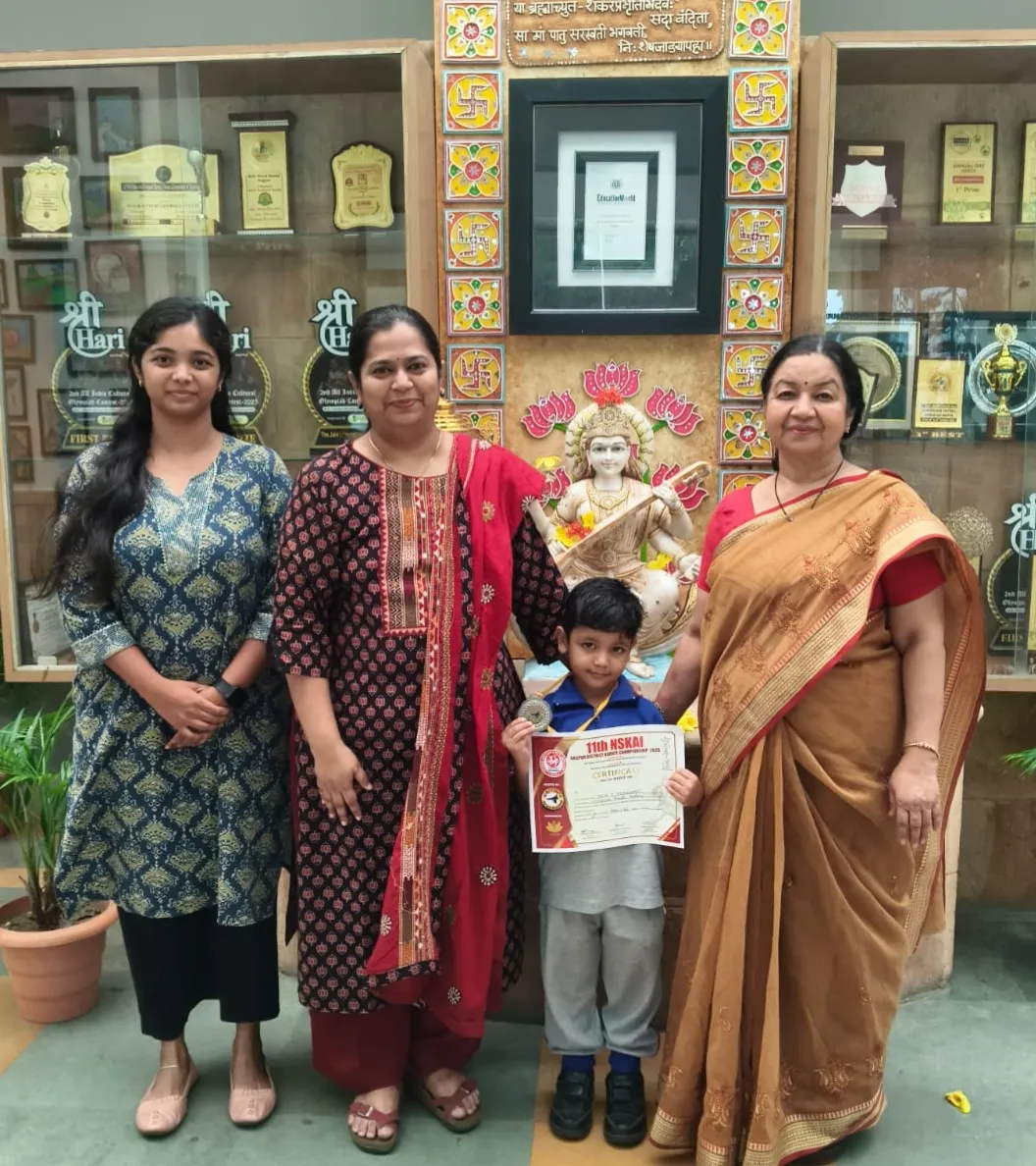 Three women and a young boy holding a certificate and medal pose in front of a Saraswati statue and trophy display cases at JIS