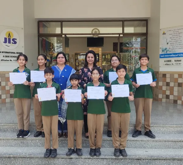 Group photo of eight students in green uniforms holding certificates, standing on steps with two women teachers in front of Jain International school entrance.