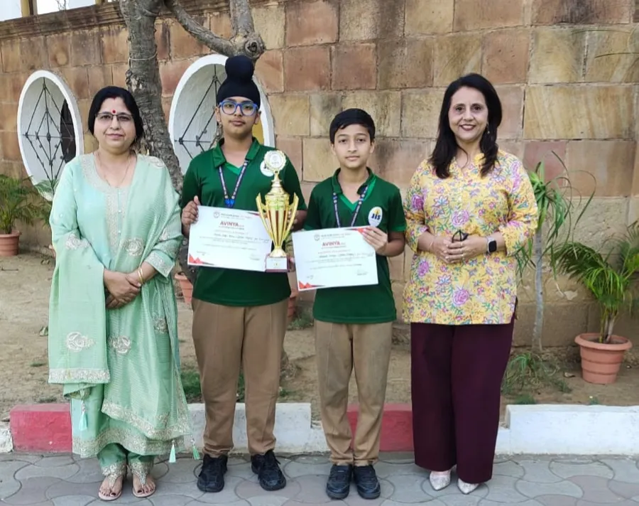 Two students in green uniforms hold certificates and a trophy, standing outdoors between two women in front of a stone wall.