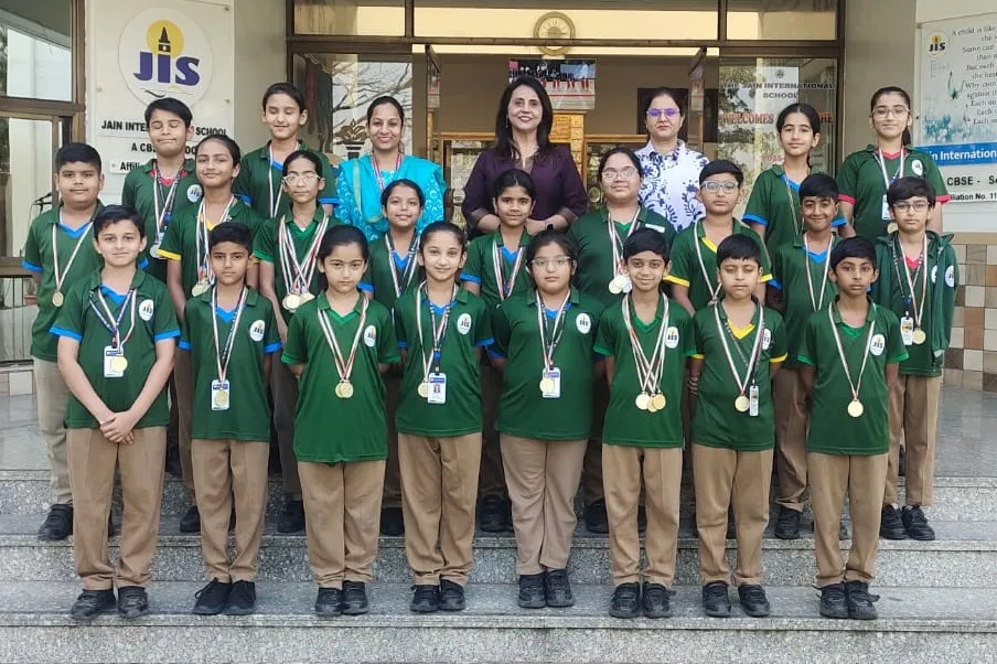 Group photo of about 20 students in green uniforms wearing medals, posed on steps with four adult women behind them in front of Jain International school entrance.