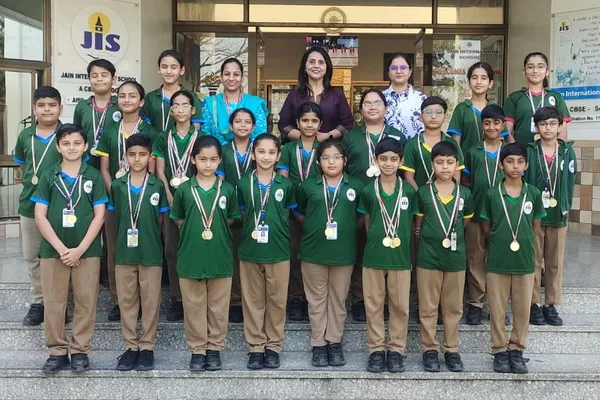 Group photo of about 20 students in green uniforms wearing medals, posed on steps with four adult women behind them in front of Jain International school entrance.