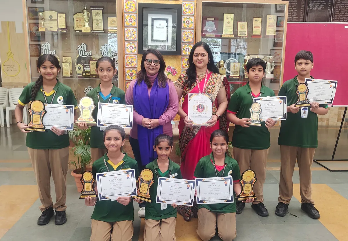 Group photo of seven students in green uniforms and two women holding certificates and trophies, standing in front of a trophy display cabinet at Jain International.