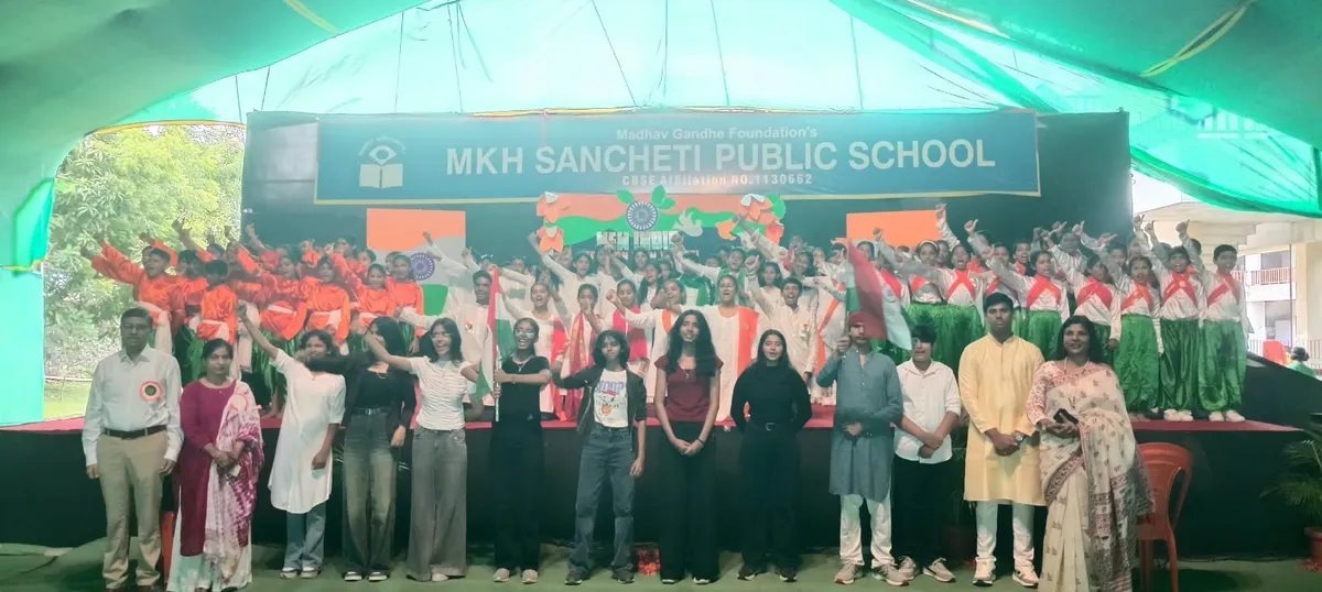 Group photo on a stage under a tent of many students in orange, white and green costumes with adults and young people in front, beneath MKH Sancheti banner.