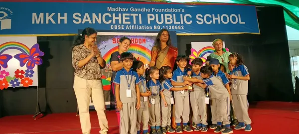 Group of young children in school uniforms on stage with four adult women, MKH Sancheti school banner and colorful decorations behind them.