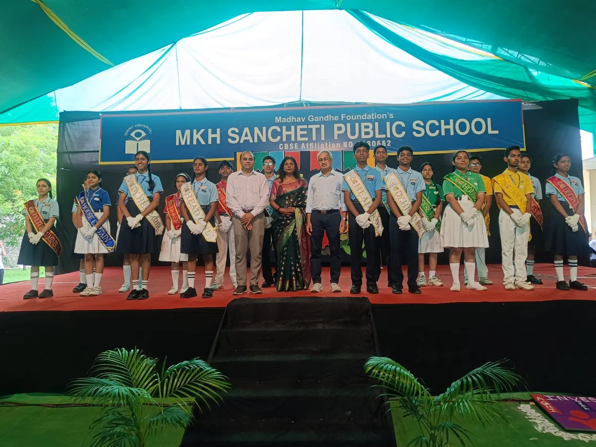 Group photo on stage of students wearing sashes standing with several adults under a tent, with MKH Sancheti school name banner behind them.