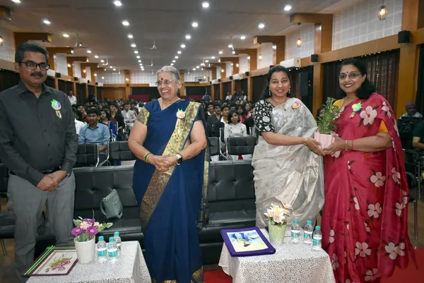 Four adults standing in front of a seated audience, one woman handing a potted plant to another at Narayana Vidyalayam.