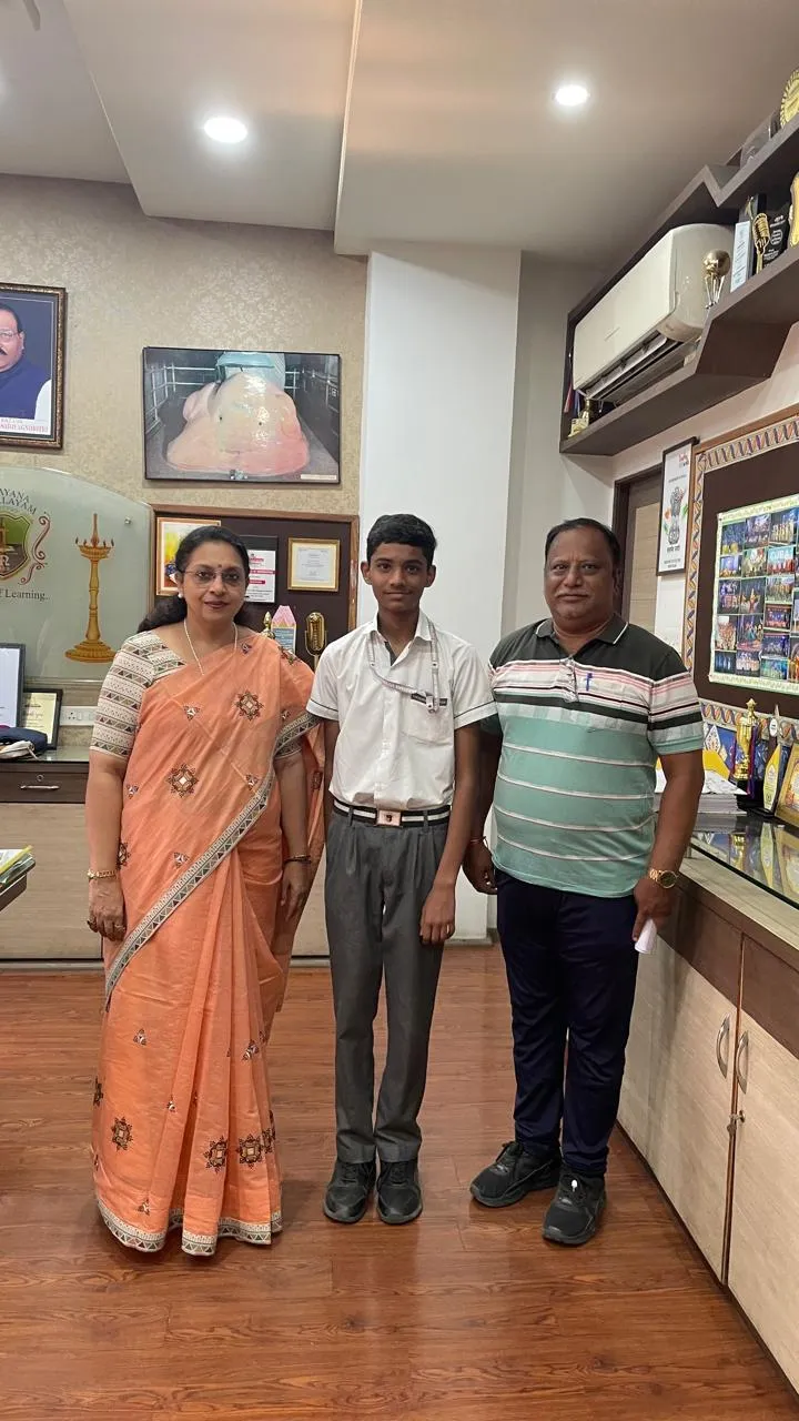 A boy in school uniform stands between a woman in an orange saree and a man in a striped shirt inside an office with trophies and framed pictures. - Narayana Vidyalayam