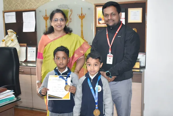 Two young boys wearing medals and holding a certificate pose indoors with a woman in a yellow saree and a man in a dark jacket.