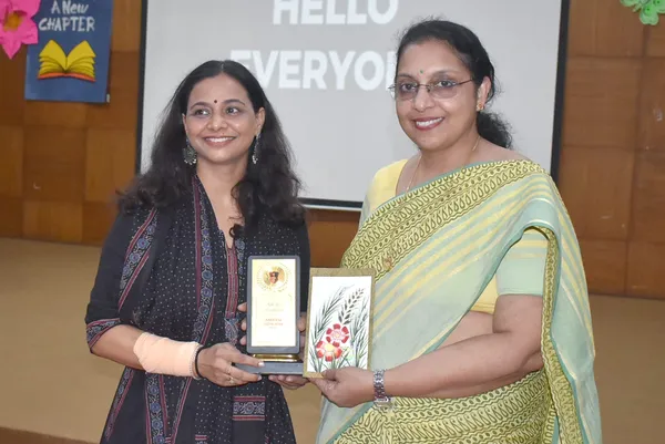 Two smiling women on a stage, one in a dark printed outfit and one in a green saree, holding a small plaque and a painted floral card.