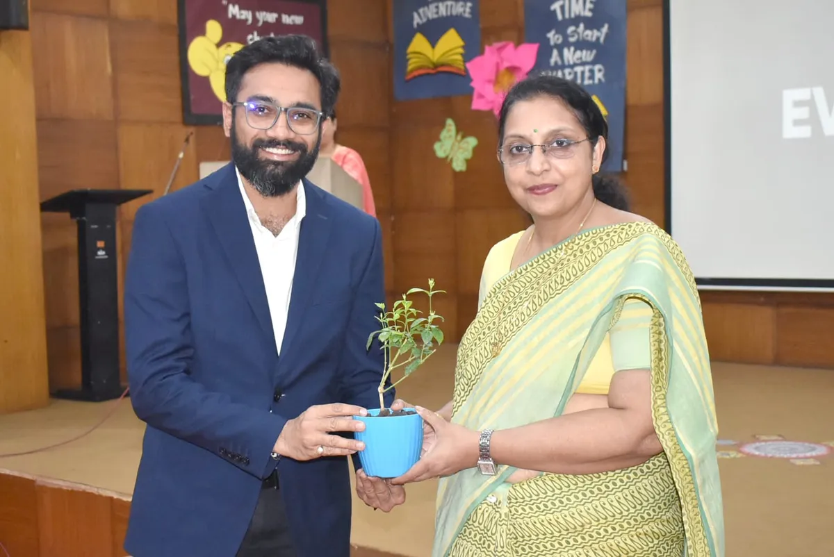 A bearded man in a blue blazer and a woman in a green saree jointly hold a small potted plant on a stage with decorated boards behind them.