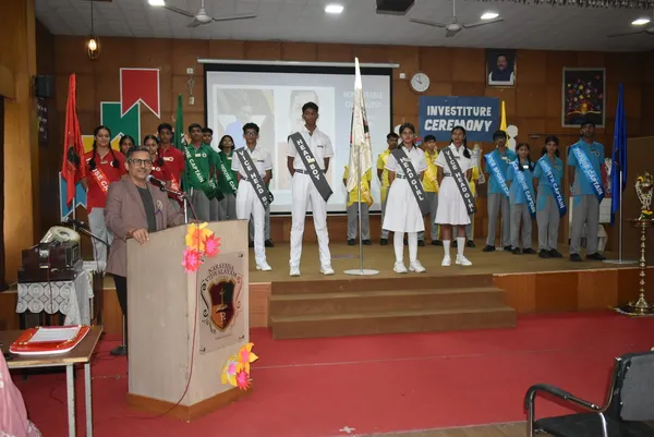 A man speaks at a podium while student leaders in sashes and house colours stand on a stage at an investiture ceremony at Narayana Vidyalayam.