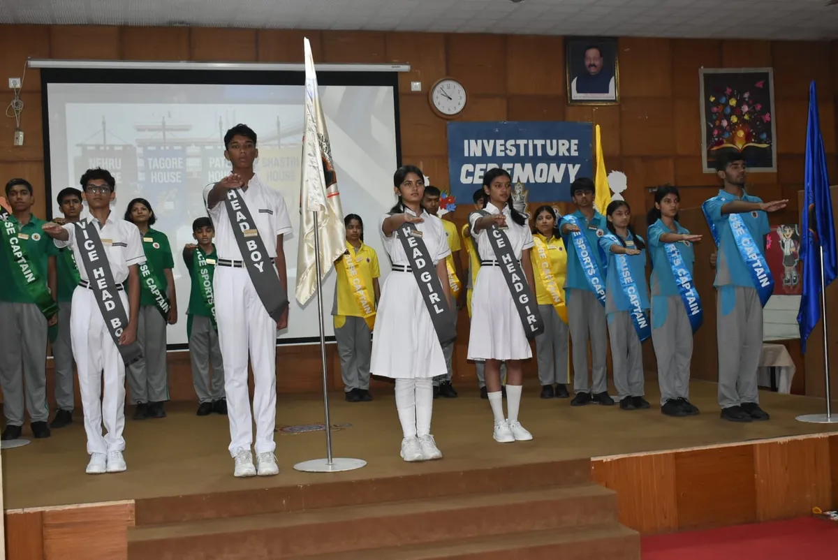 Students wearing sashes stand on a stage with raised hands taking an oath at an investiture ceremony, with a flag and projector screen behind them at Narayana Vidyalayam.