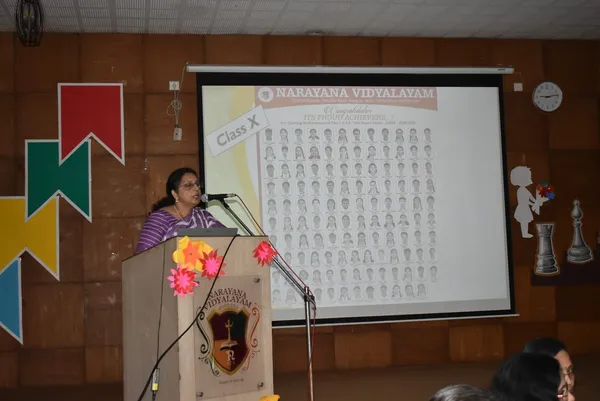 A woman speaking at a decorated podium beside a projected slide showing a grid of student portraits, with audience heads visible in the foreground