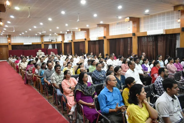 A large audience of adults seated in rows in a hall with a red carpet aisle