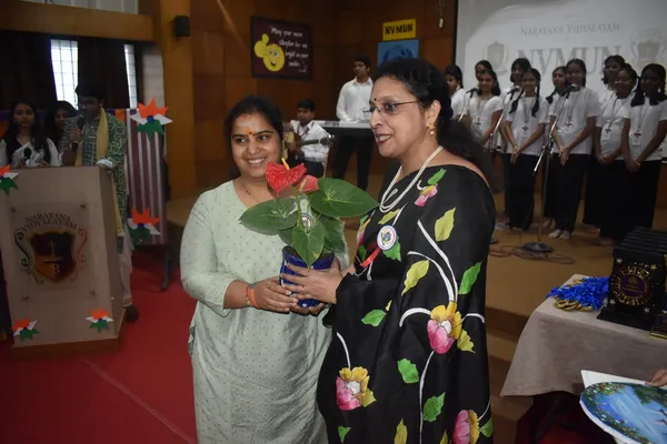 Two women hold a potted red anthurium plant on a stage at Narayana Vidyalayam, with students standing in rows behind them and a podium and trophies nearby.