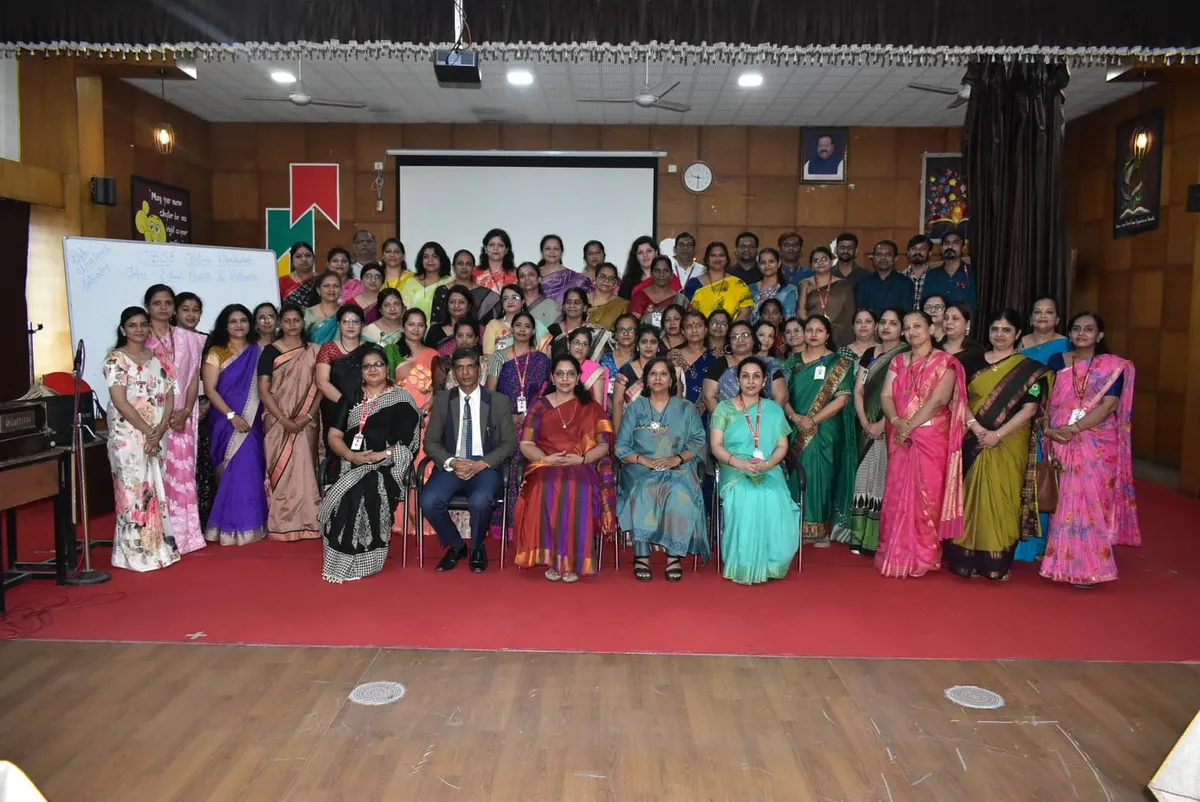 Large group photo of teachers, mostly women in colourful sarees, posed in rows on a red carpet on a hall stage with a screen and whiteboard behind them at Narayana Vidyalayam.